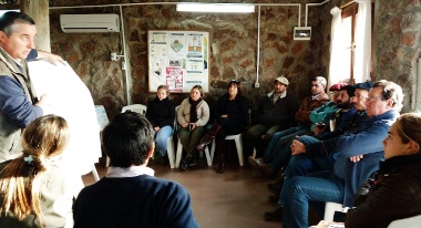 Grupo de familias colonas, estudiantes y técnicos dentro de un salón escuchan a Julio Perrachón dar su charla